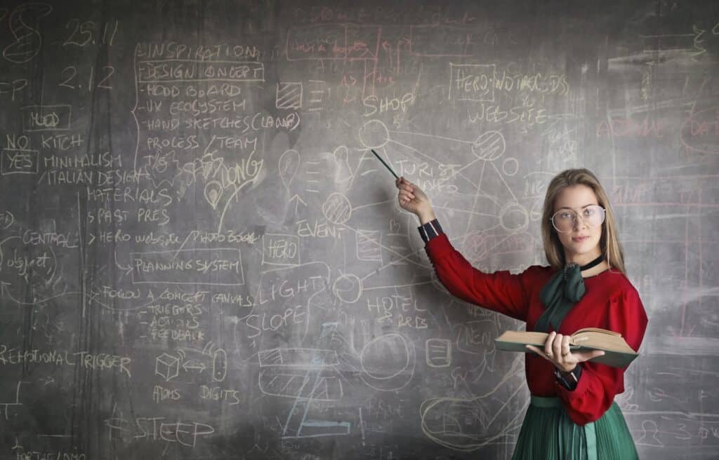 Academic success coach points at a blackboard while reading from a book held in her other hand.