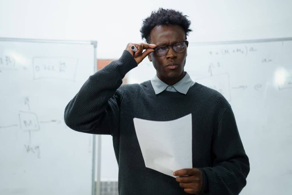 A black man stands looking quizzically at a paper he is holding while standing in front of a whiteboard. 