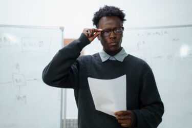 A black man stands looking quizzically at a paper he is holding while standing in front of a whiteboard. The image represents a graduate student wondering what to do to prepare for graduate school.
