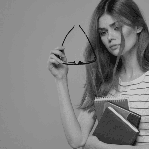 An image of a woman taking off her glasses with one hand and holding books in the other. Her expression is one of concern bordering on worry.