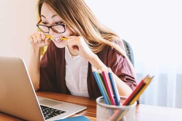 Woman sits at her computer biting her pencil. Frustration is evident in her face. Featured image shows student struggling with academic writer's block.