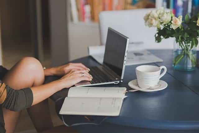 Featured image for improving scholarly writing blog post displays a cropped image of a student's hands typing on a laptop with a notebook and teacup on the table beside them.