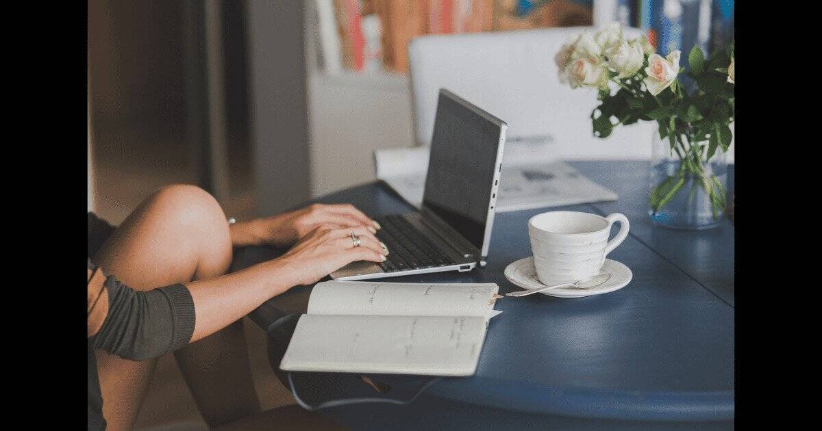 Cropped image of an academic writing on a laptop with a notebook and cup of tea beside them. A vase of flowers and open peer-reviewed journal are on the table behind the laptop.