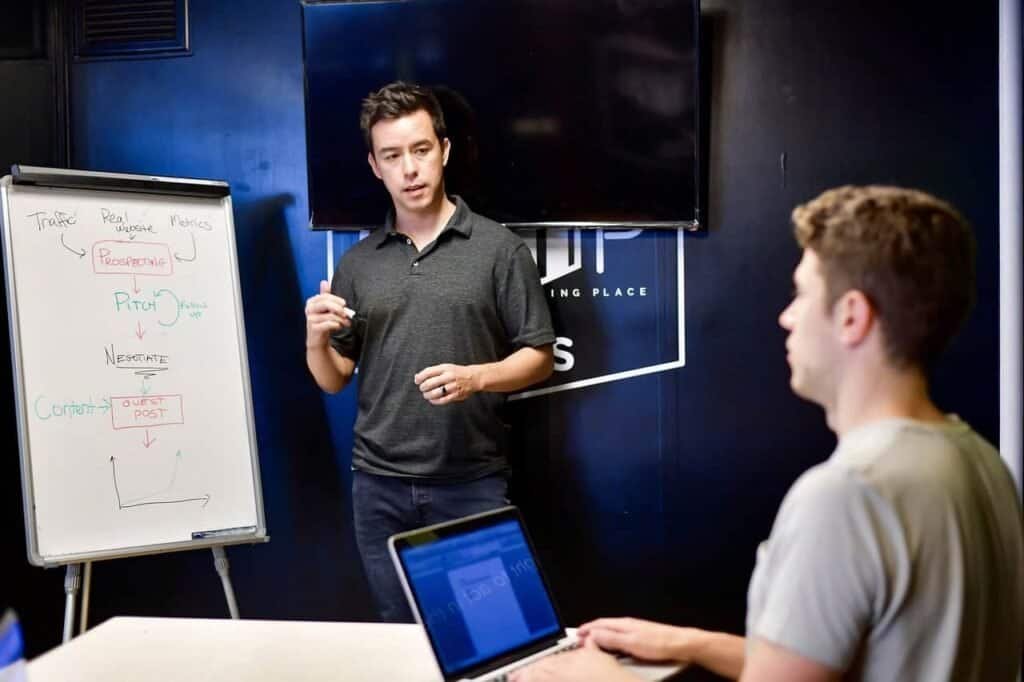 Image of a graduate student adviser standing by a white board, talking to a student who is taking notes on a laptop.