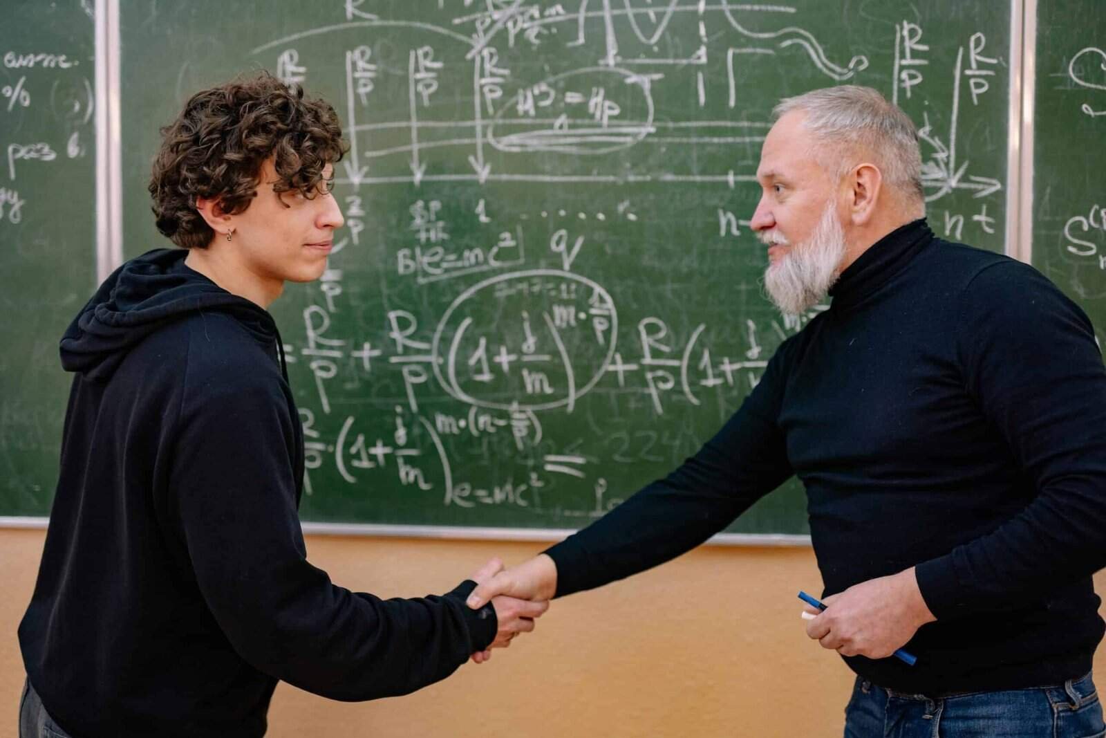 A PhD advisor shakes a prospective student's hand in front of a chalkboard.